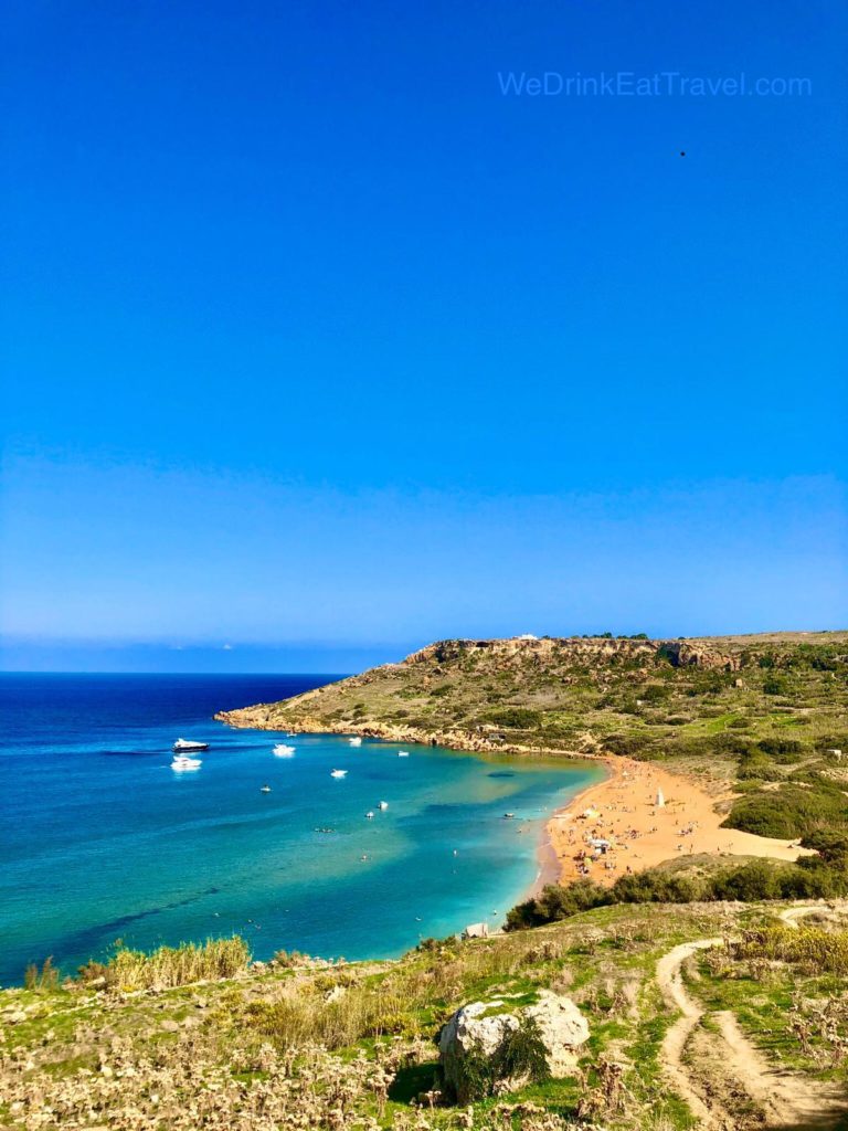 A sweeping, elevated vista looking out from the clifftop near Calypso Cave in Xagħra, Gozo, showcasing the striking red-sand beach of Ramla Bay and its surrounding green valley.