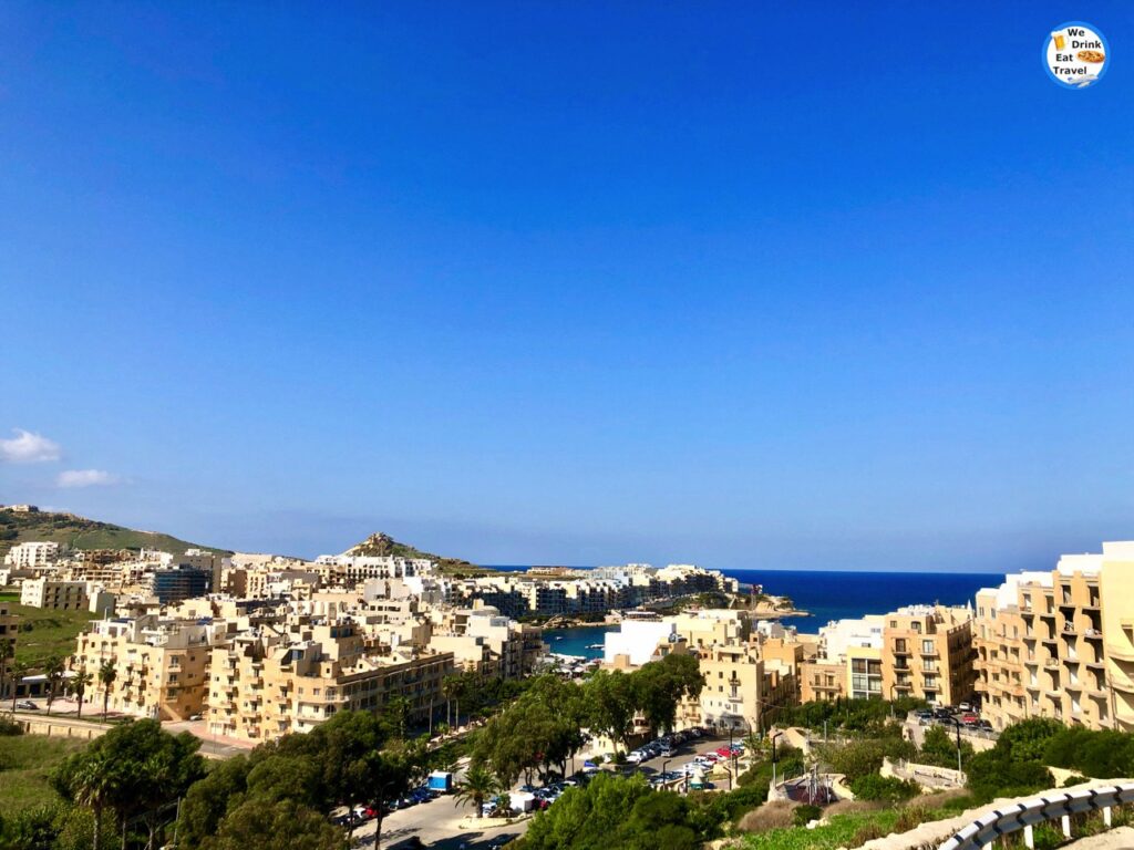 A panoramic elevated view of Marsalforn Bay in Gozo, showcasing the crescent-shaped coastline, buildings lining the waterfront, moored boats in the clear blue water, and surrounding rugged hills.