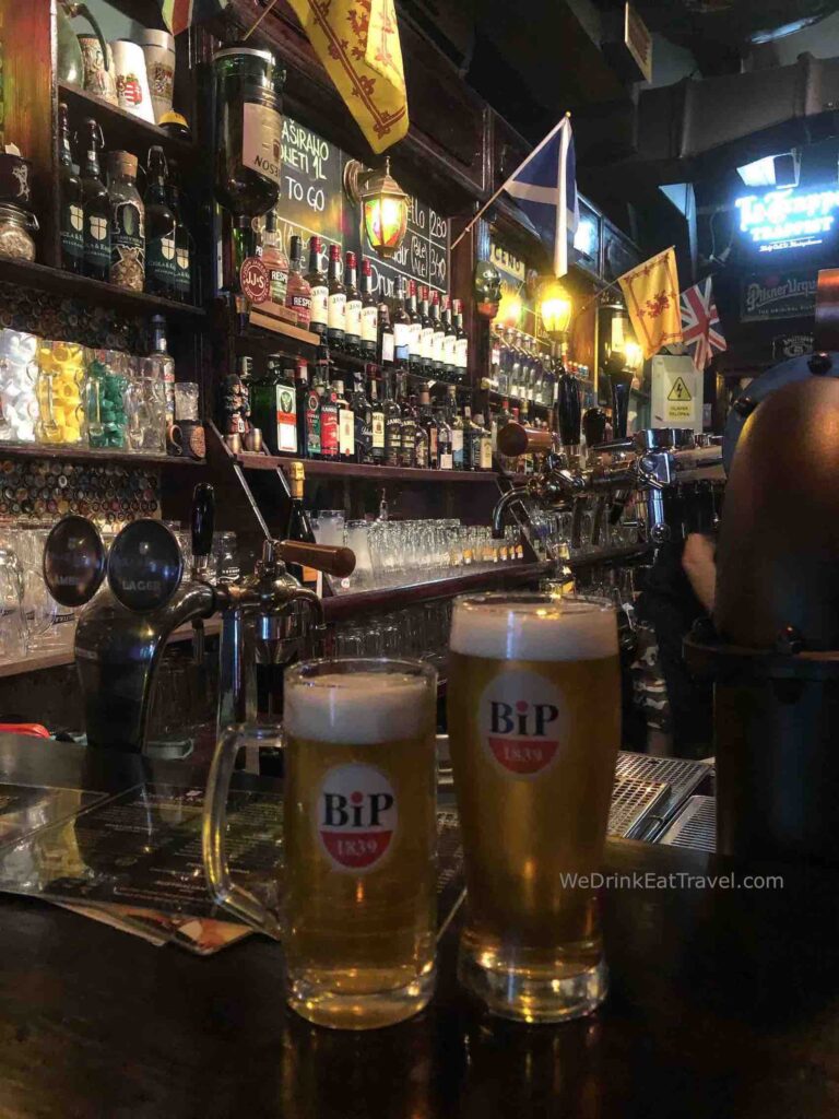 One of the top bars in Belgrade, Serbia showcasing 2 beers in a glass with the bar in the background fully stocked with glasses and a wide variety of spirits.