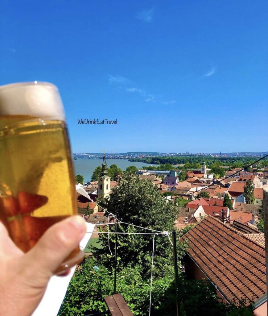 Person holding up a beer, with terracotta color rooftops, lush green landscape and river at the landscape background with a brilliant clear blue sky.