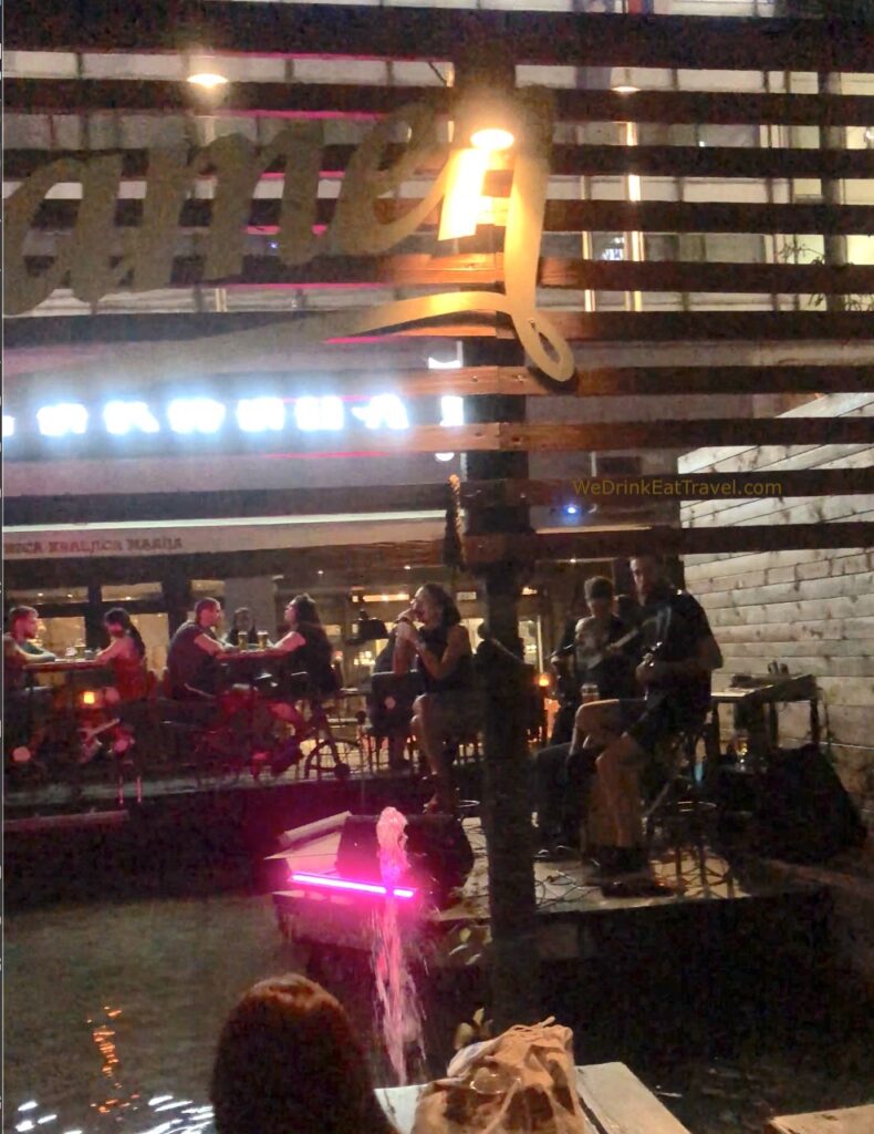 A band and their singer on a small platoon-like stage above water at one of the bars in Belgrade, surrounded by people drinking at tables and chairs.