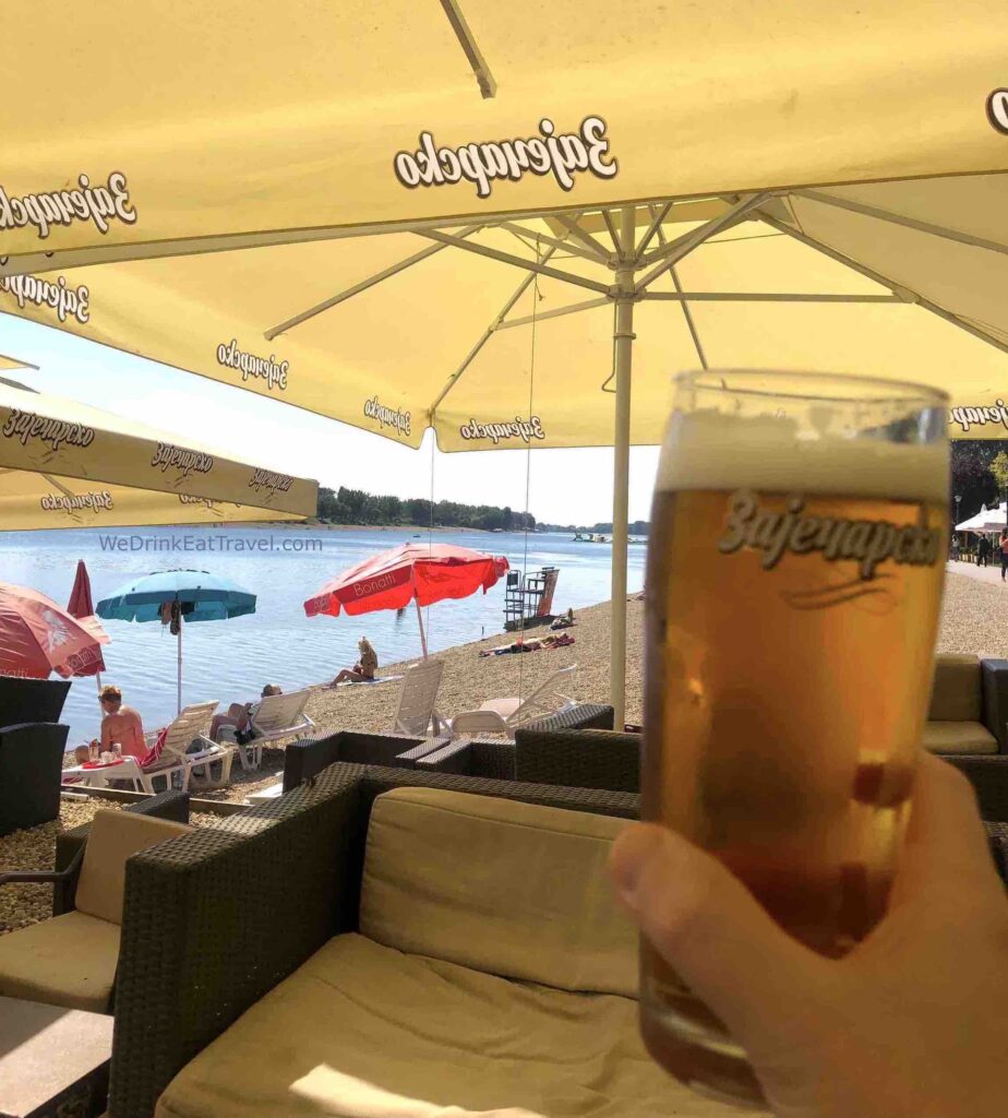 A big glass of beer being held up with people lying on a rocky beach and sun loungers under colourful umbrellas in Belgrade, Serbia.