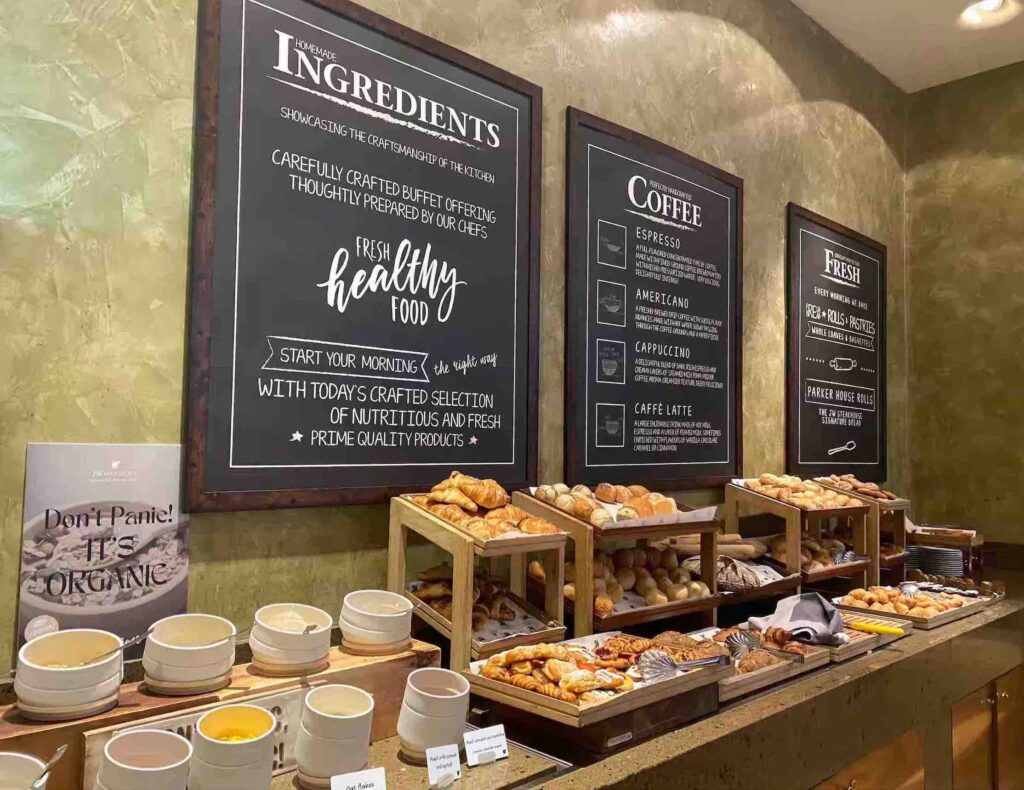 A large selection of breads, pastries, danishes and condiments on a long table with 3 blackboards behind it mentioning the freshness and quality of food and drinks at the JW Steakhouse restaurant, Bucharest.