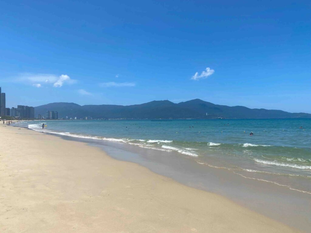 Where to stay in Da Nang? Here is a beautiful view of the white sand, turquoise water and mountain range at My An Beach, a beautiful Danang beach location.