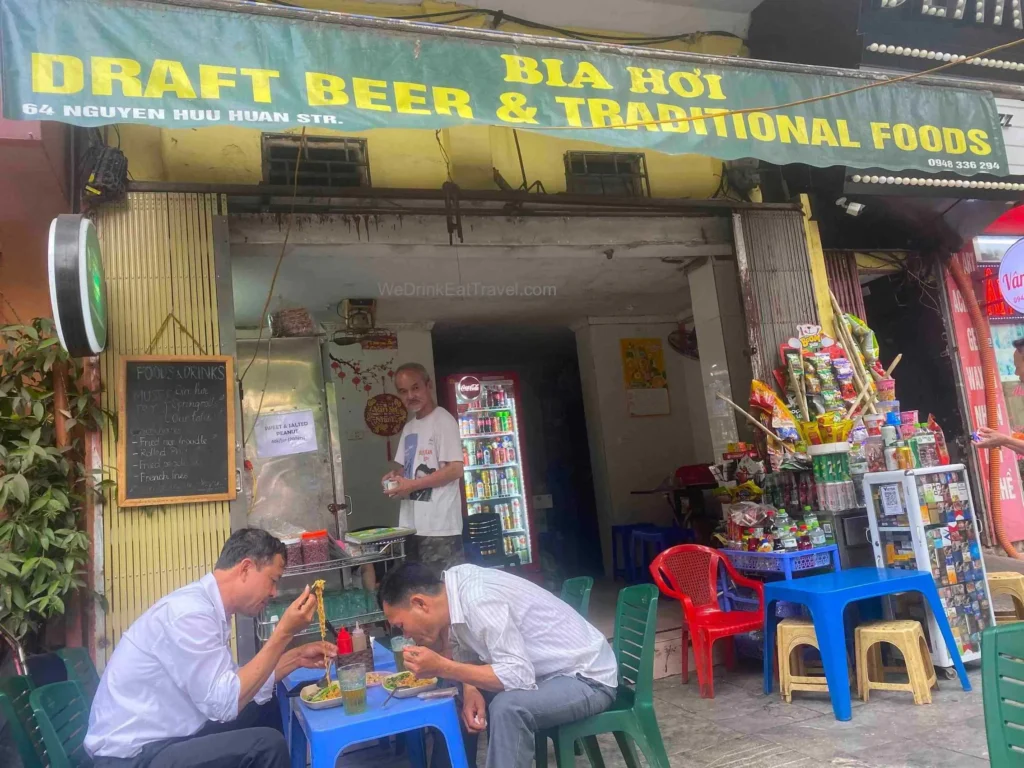 Two men eat and drink bia hoi on small blue plastic chairs at the front of HaNoi Draftbeer & Traditional food in Hanoi Old Quarter.
