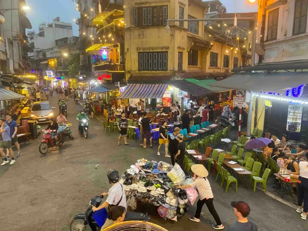 Overlooking the famous bar street in Hanoi Old Quarter at dusk, with colorful lanterns, outdoor seating, motorbikes, and vendors selling shoes and food.