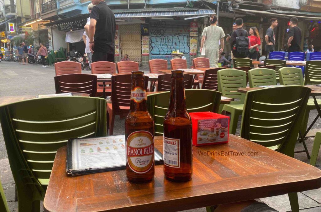 Two bottles of Hanoi Beer on a wooden table surrounded by green and brown plastic chairs with people roaming the streets in Hanoi Old Quarter.