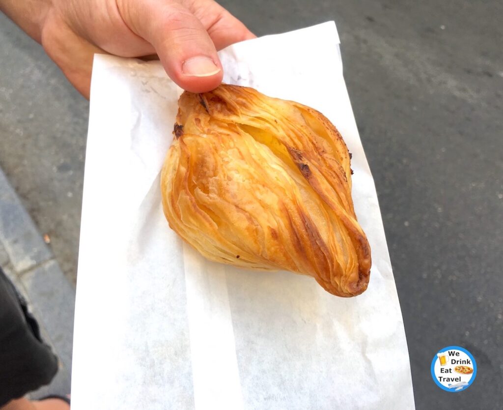 One of the best pastizzi in Malta with a close-up of a ricotta pastizzi, with layers of golden flaky pastry being held on a white paper bag.