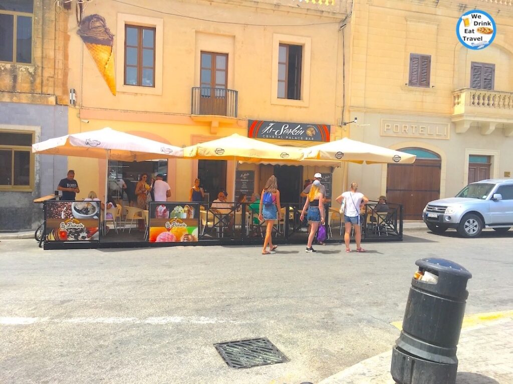 Storefront of a popular restaurant in Malta with customers sitting out front under big umbrellas with bright sunshine glowing.
