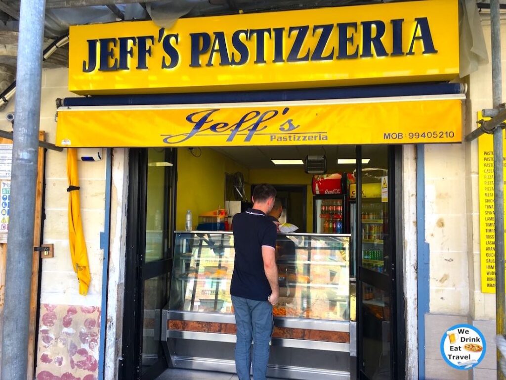 Open shopfront for Jeff's Pastizzeria in Malta with bright yellow and navy blue signage, with a customer buying food in front of a glass display cabinet.