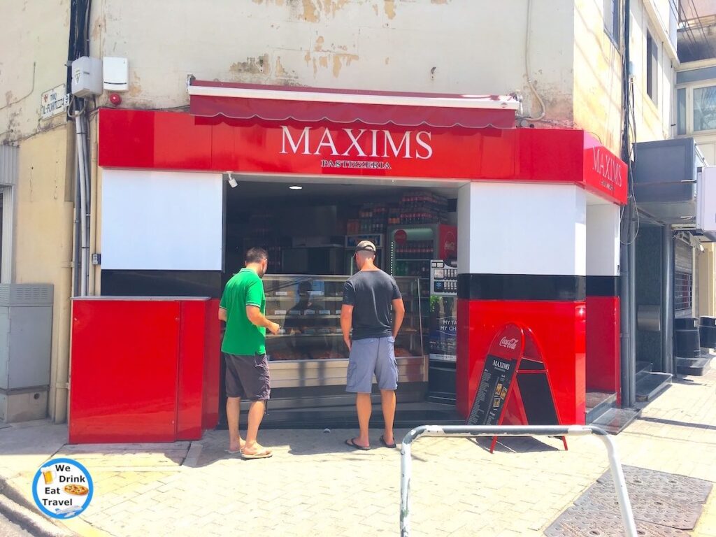 Looking for the best pastizzi in Malta with customers looking at food in front of a big glass display cabinet, and an A-frame showing menu items at the shop front.