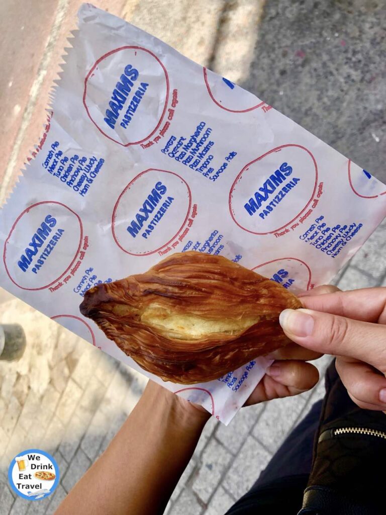 Delicious Malta food showcasing a close-up of a ricotta pastizzi, with layers of deep golden flaky pastry being held on a branded white paper bag.