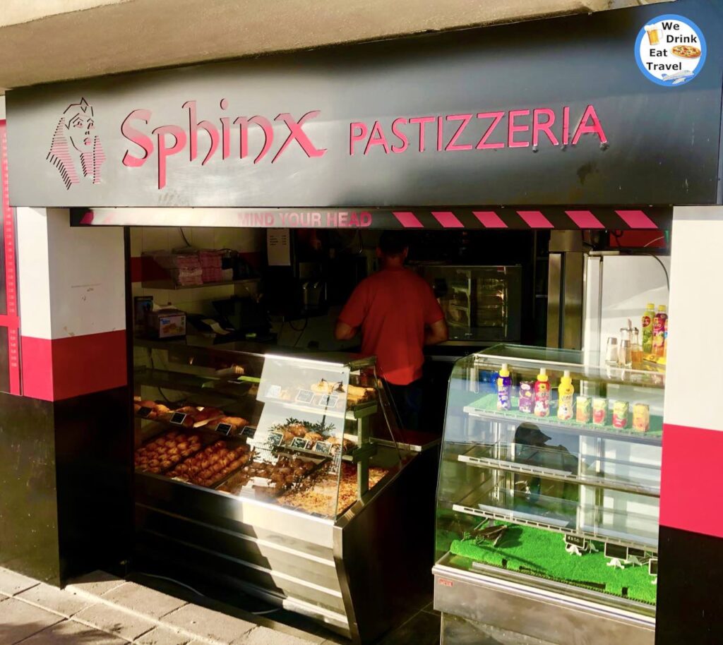 Open shopfront for Sphinx Pastizzeria with fluorescent pink signage, showcasing a variety of Malta food and drinks in glass display cabinets.