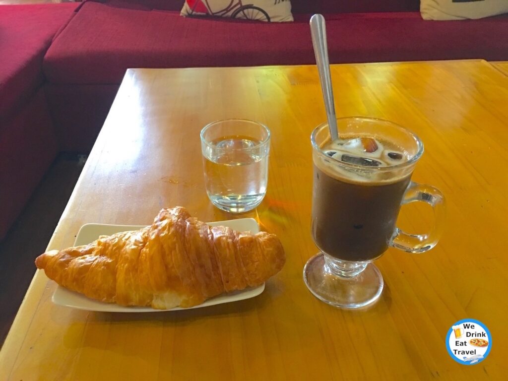 Butter croissant on a white plate and Vietnamese iced coffee with condensed milk on a table, with maroon couches surrounding them.