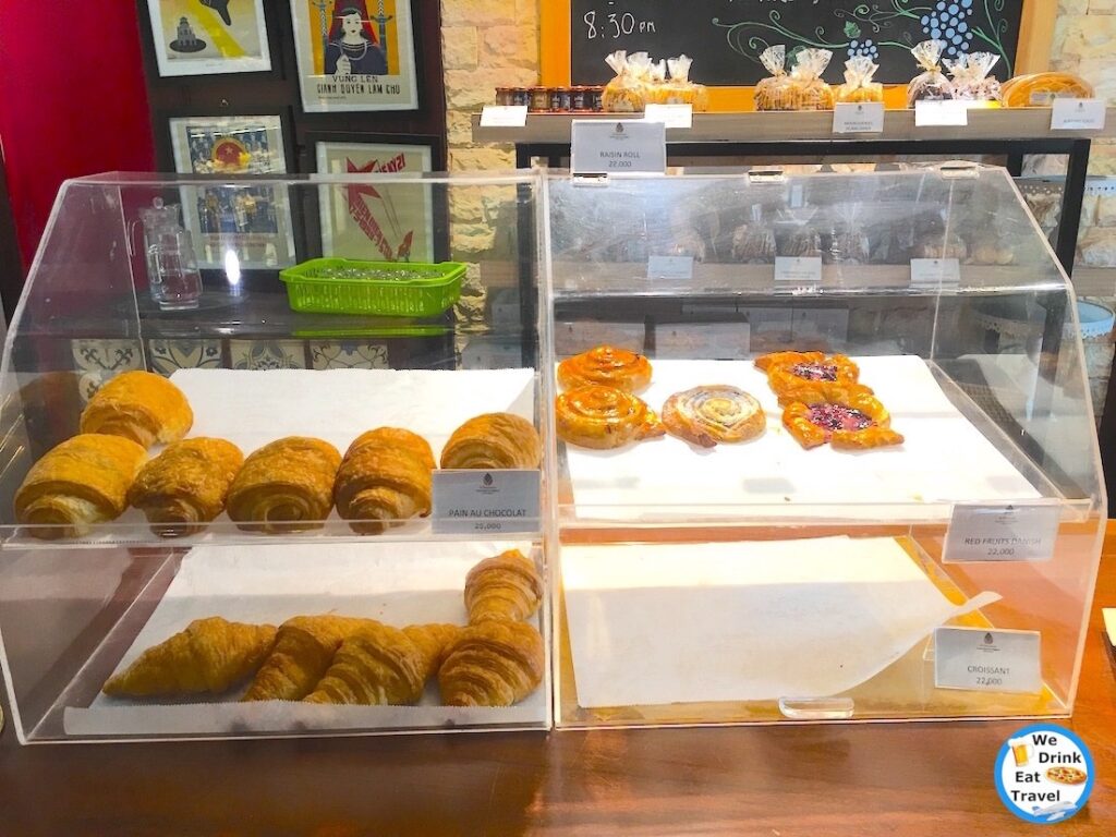 Pastries on display in a clear cabinet at a French Bakery in Hanoi Vietnam, with pictures hanging behind, and jams and cookies for sale in the background.