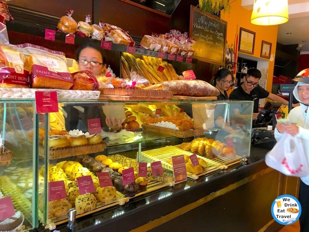 Pastries, cakes and breads on display in a clear cabinets and wooden shelves at a French Bakery in Hanoi Vietnam, with staff serving customers.
