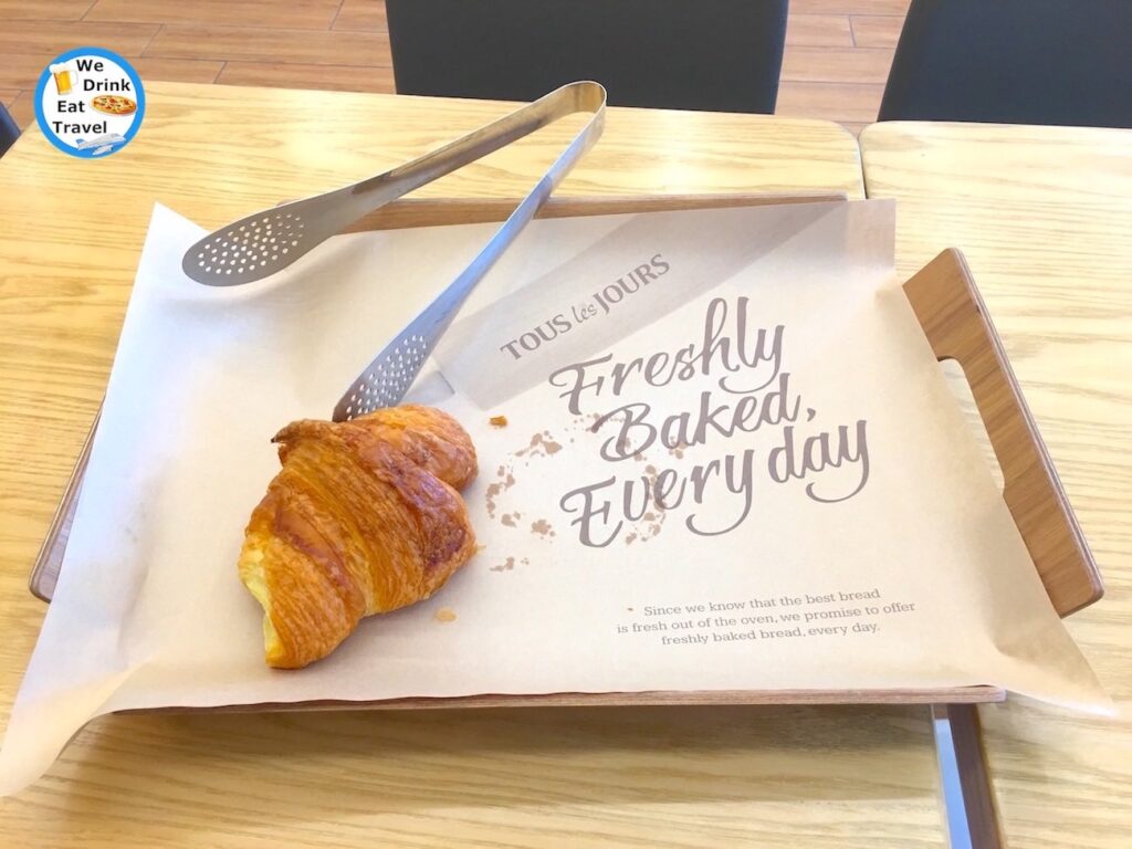 Close-up of a fresh butter croissant in a Tous Les Jours French Bakery in Hanoi Vietnam, served on a wooden tray with tongs, and a branded placement with promotional wording.