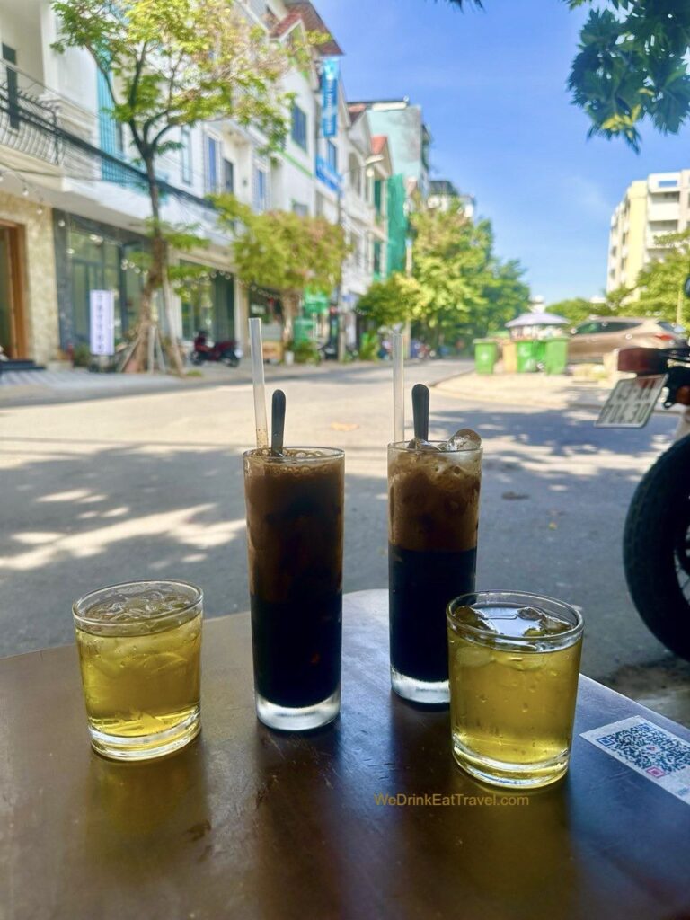 Two tall glasses of Vietnamese black coffee, ca phe den da, with two small glasses filled with tea. Buildings, green leafy trees and a bright clear blue sky in the background.