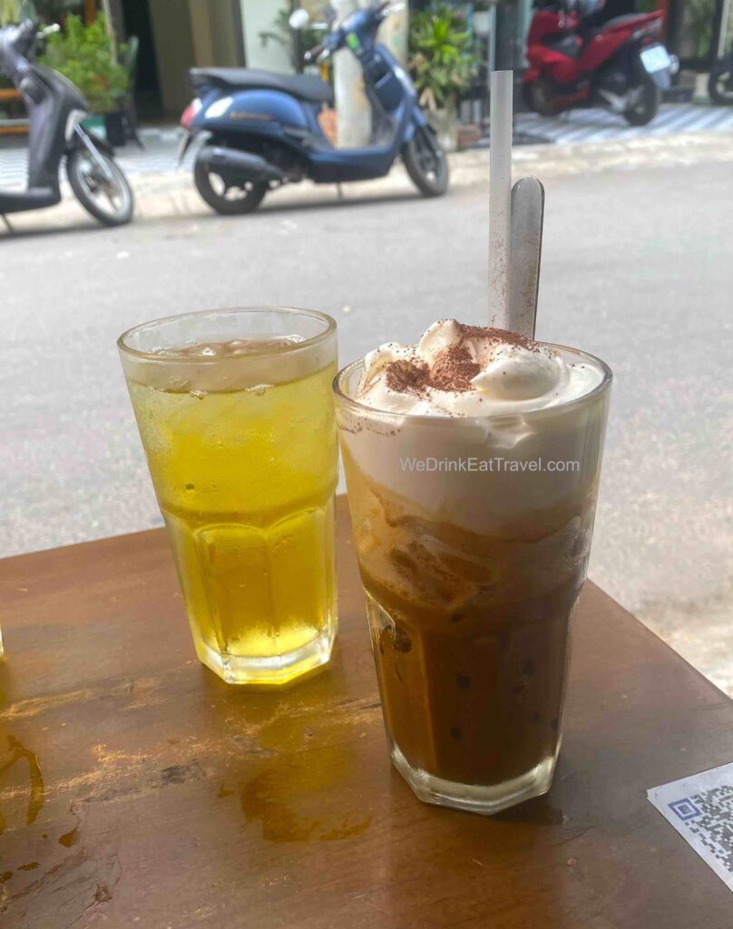 Close-up of a Vietnamese salt coffee, ca phe muoi with the rich dark coffee at the bottom, topped with a salt cream layer and hint of powdered chocolate, accompanied by a glass of tea.