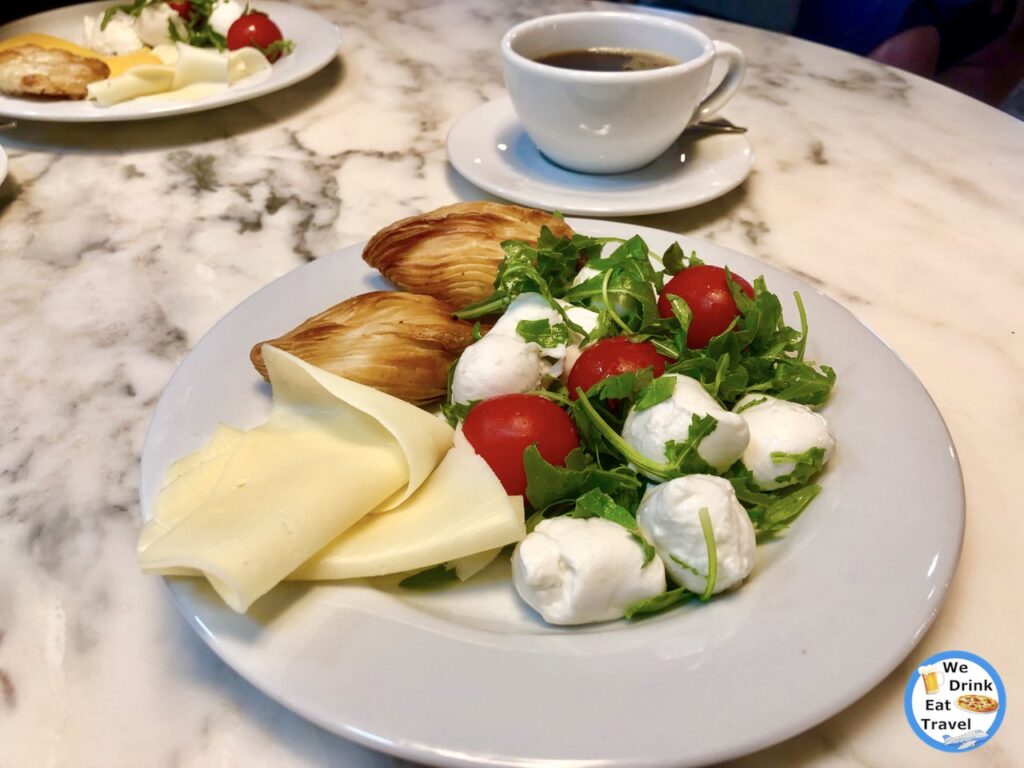 Assorted cheeses, cherry tomatoes, rocket and flaky mini pastizzis on a white plate with a cup of black coffee in Sliema, Malta.