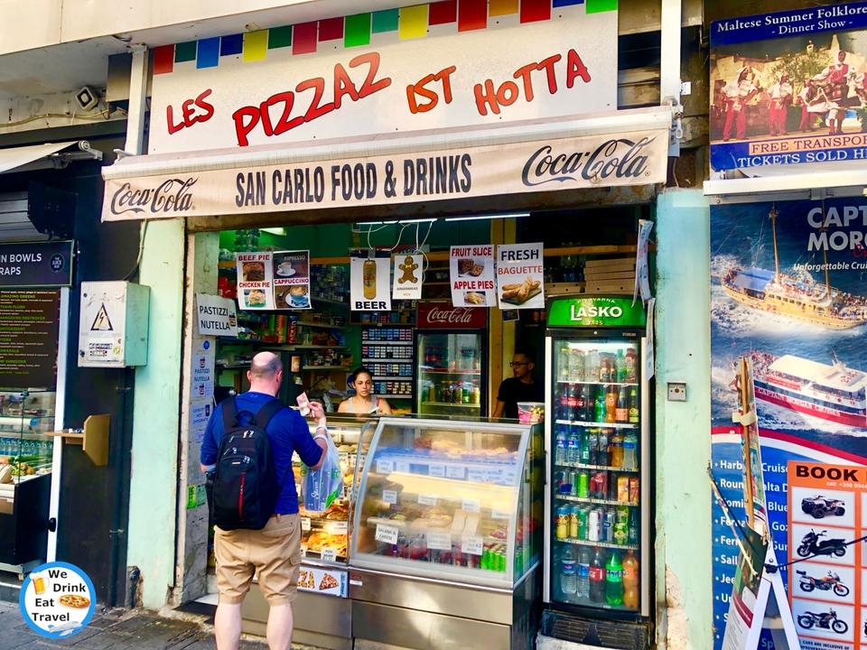Open shopfront for a take away store in Sliema, Malta showcasing a variety of food and drinks in glass display cabinets and signs with pictures showing different food and drinks on offer.