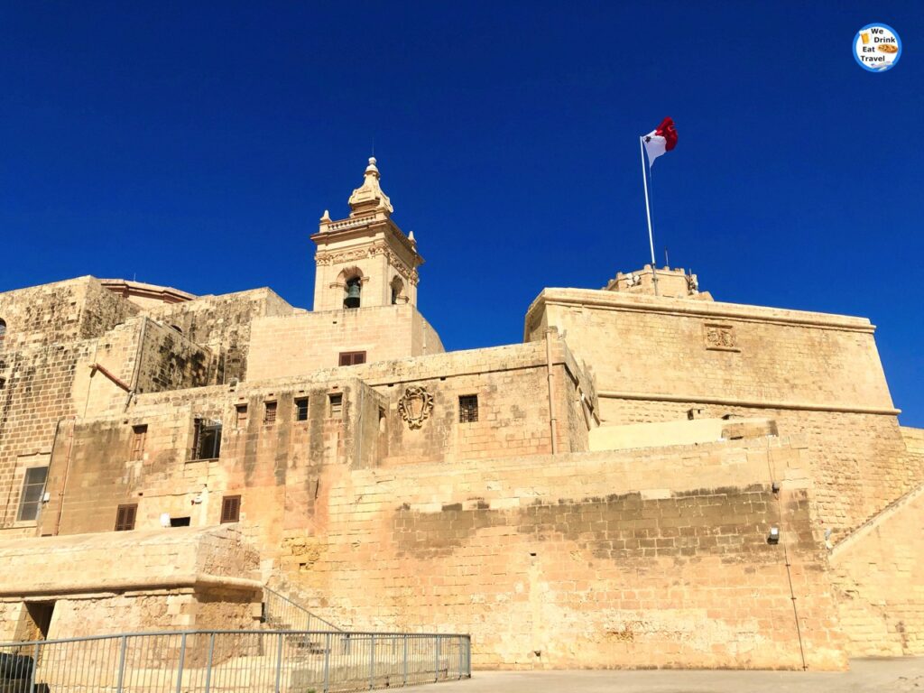 An elevated view of the ancient Cittadella fortress in Victoria, Gozo, showcasing its honey-colored stone bastions, church bell and Maltese flag under a bright Cleary blue sky.