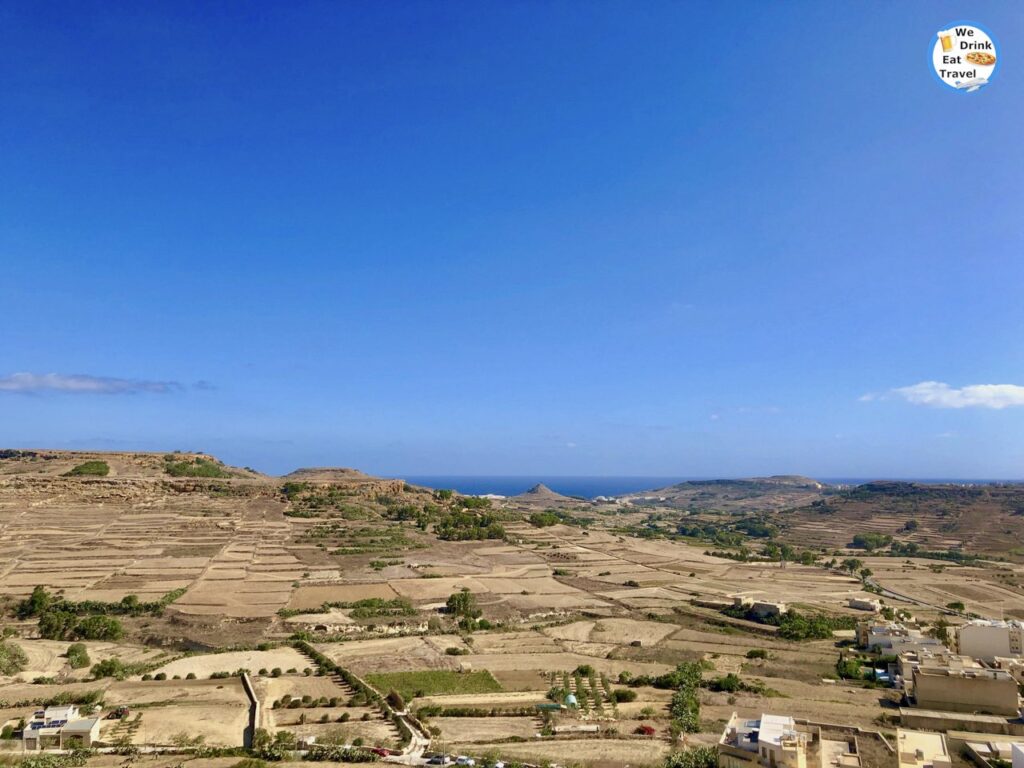 View looking out from the historic fortifications of the Cittadella in Gozo, showing a patchwork of green and beige agricultural fields, scattered buildings, and rolling hills under a bright sky.