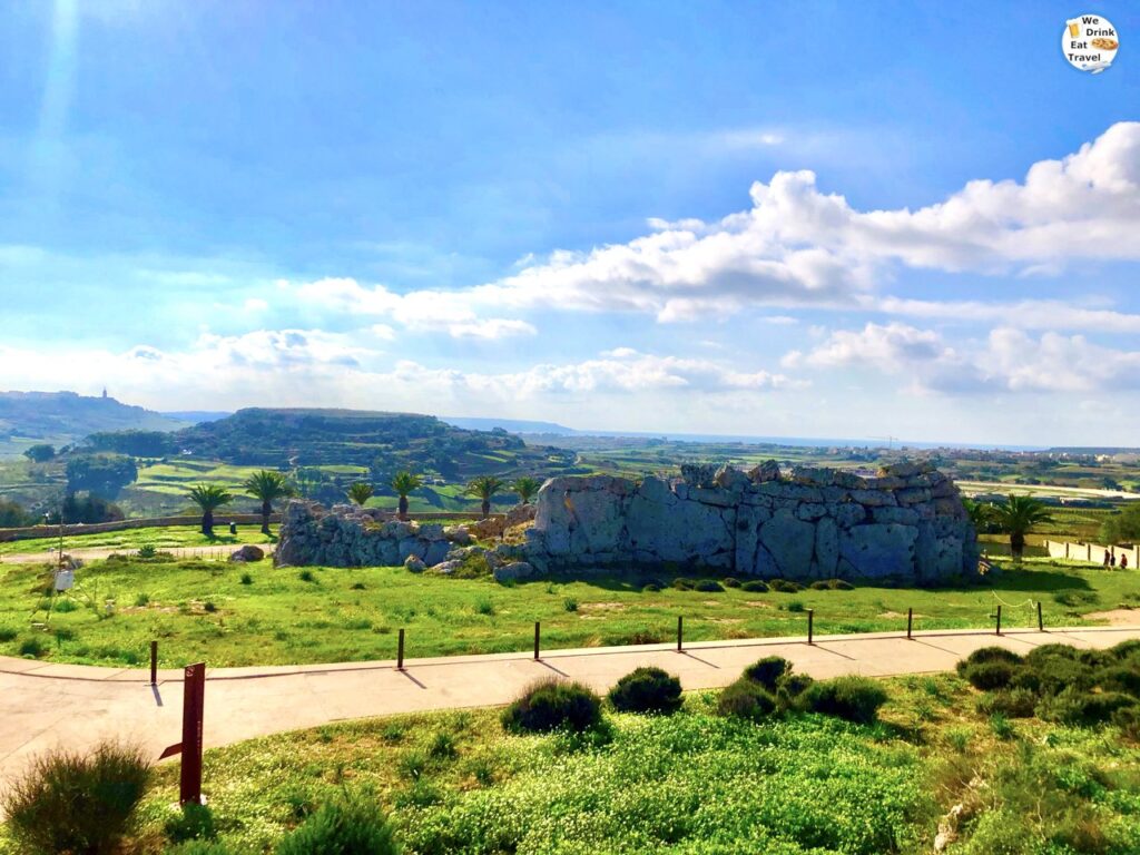 A wide-angle view of the prehistoric Ġgantija archaeological site in Gozo, showcasing massive, weathered megalithic limestone blocks forming the ancient temple walls under a bright sky.
