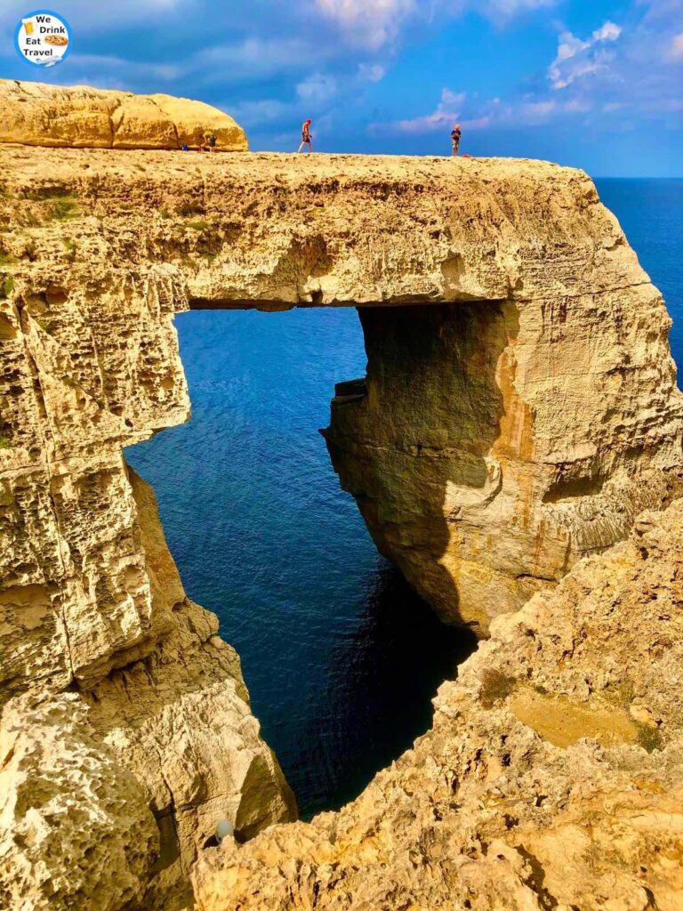 Close up of the Ghasri Valley (Wied il-Ghasri) in Gozo, Malta with the golden rock window and clear blue waters surrounding it under a bright blue sky with a few powdery white clouds.