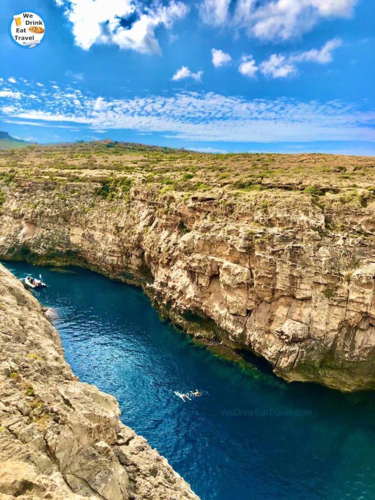 Ghasri Valley (Wied il-Ghasri) in Gozo, Malta with 2 people swimming in the deep blue water with rock formations on either side, and a view of the green landscape.