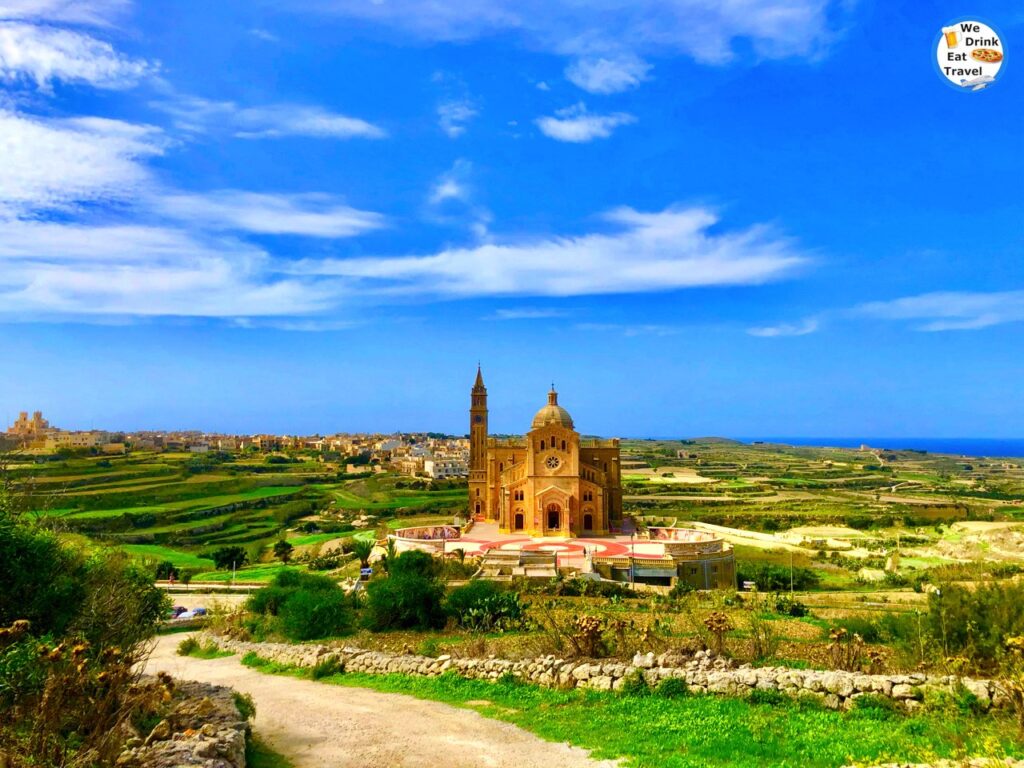 A wide-angle landscape view featuring the ornate architecture of Ta Pinu standing proudly in the center, surrounded by tranquil, terraced fields and the township of Għarb, Gozo, under a bright sky.