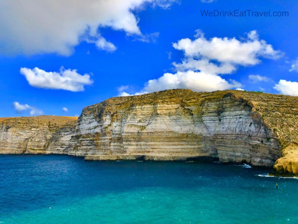 Sunlit, rugged limestone cliffs and rock formations towering over the turquoise and deep blue Mediterranean waters of Xlendi Bay on the island of Gozo, Malta.