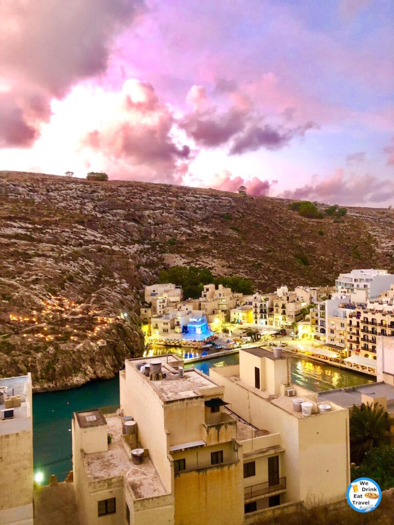 A dramatic evening sky in Xlendi, Gozo, filled with rich purple and pink hues, over urban buildings and rolling hills in the background.