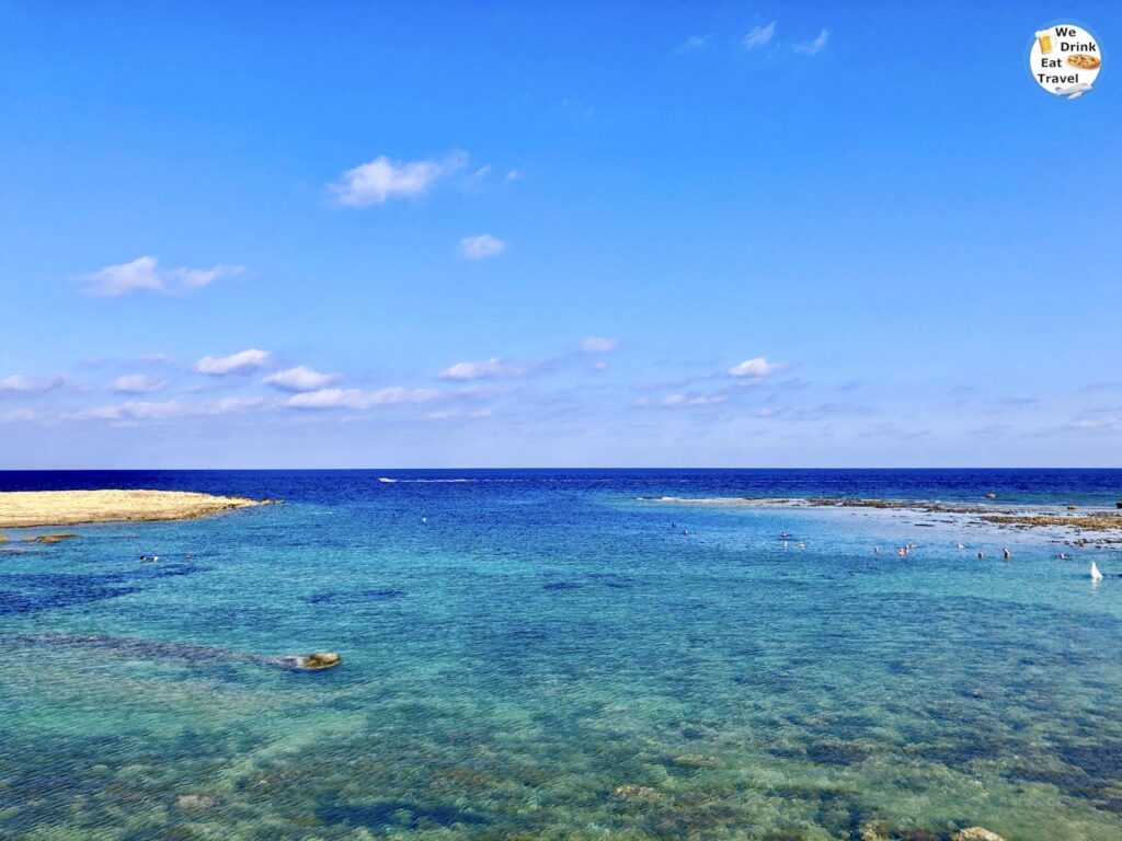 A scenic beachfront perspective of Xwejni Bay in Gozo, featuring textured rock formations along the shore, and the wide expanse of the blue sea under a bright sky.
