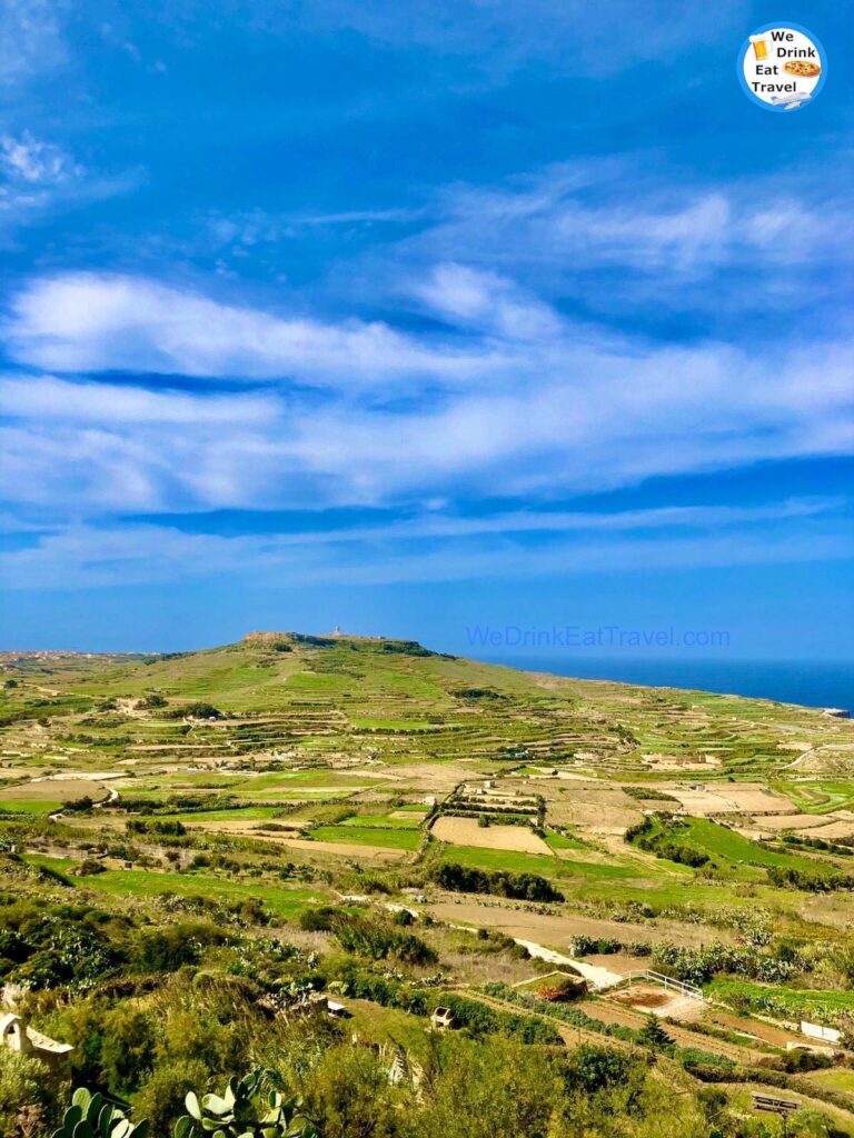 Terraced fields and lush green hills in Zebbug, Gozo, Malta, stretch towards a distant hilltop village under a vibrant blue sky with wispy clouds. The landscape evokes a sense of rural tranquility and natural beauty.
