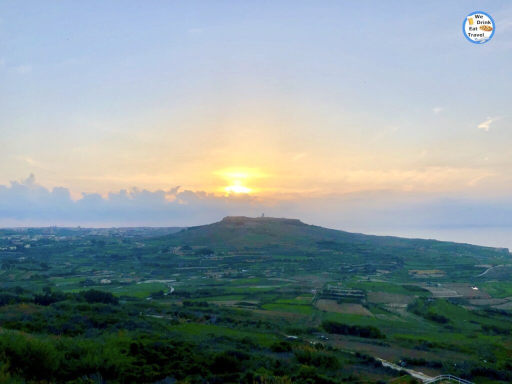 Terraced fields and lush green hills in Zebbug, Gozo, stretch towards a distant hilltop village under a colourful sunset. The landscape evokes a sense of rural tranquility and natural beauty at dusk.