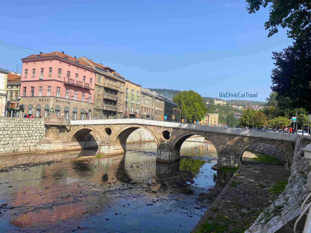 The historic Latin Bridge over the Miljacka River in Sarajevo, Bosnia and Herzegovina, with historic buildings and apartment blocks lining the banks under a clear sky.