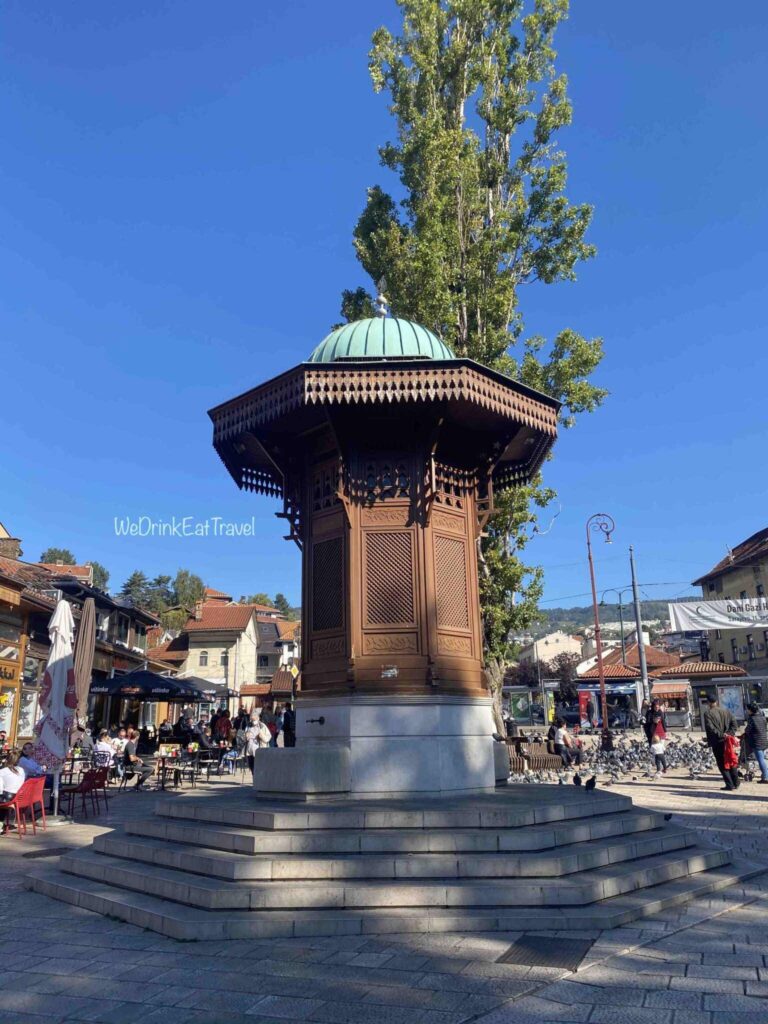 The wooden and stone fountain of Sebilj Sarajevo standing tall in the centre of a public square with pigeons and people in the background on a bright sunny day with clear blue sky.
