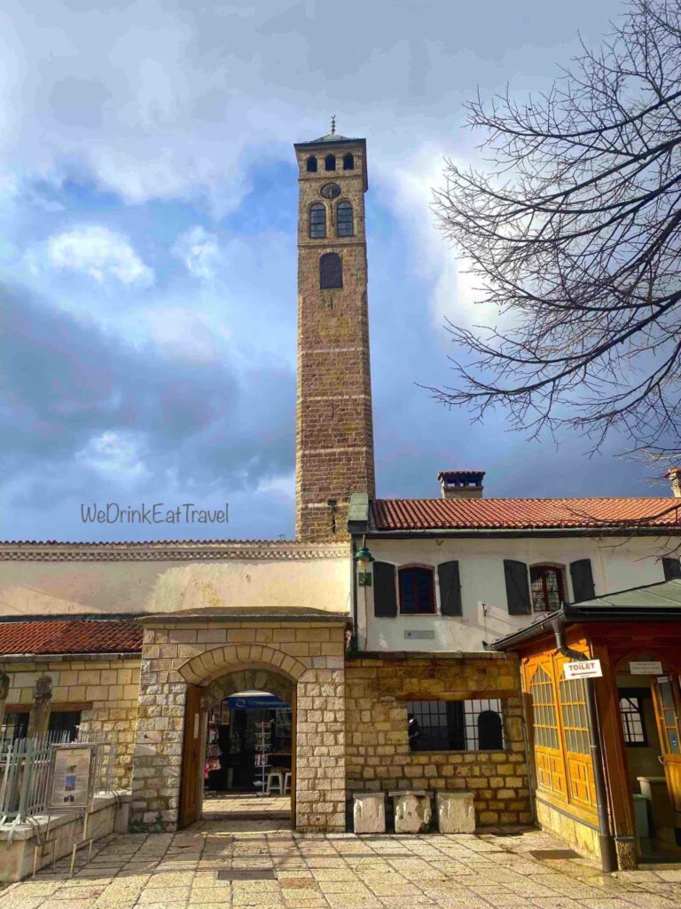 The historic tall stone, Sarajevo Clock Tower with a distinct gabled roof and arched windows, rising above the rooftops of Sarajevo's old town.