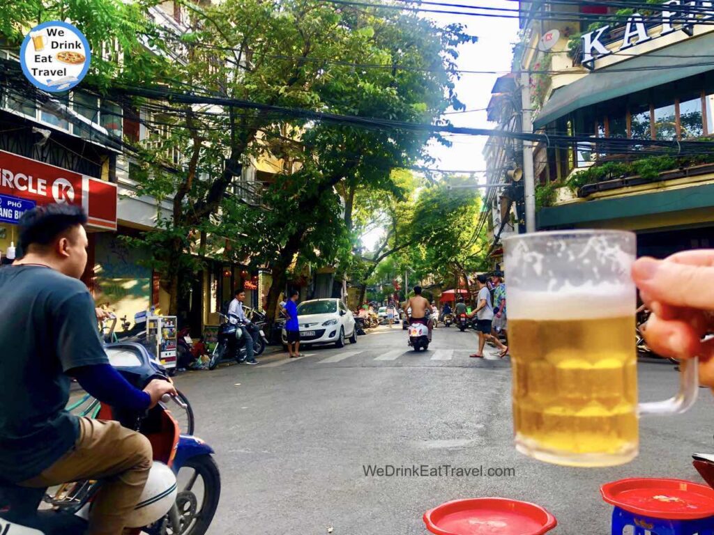Holding up a glass of bia hoi traditional Vietnamese beer in Hanoi Old Quarter with people riding motorbikes and walking the streets in the background.