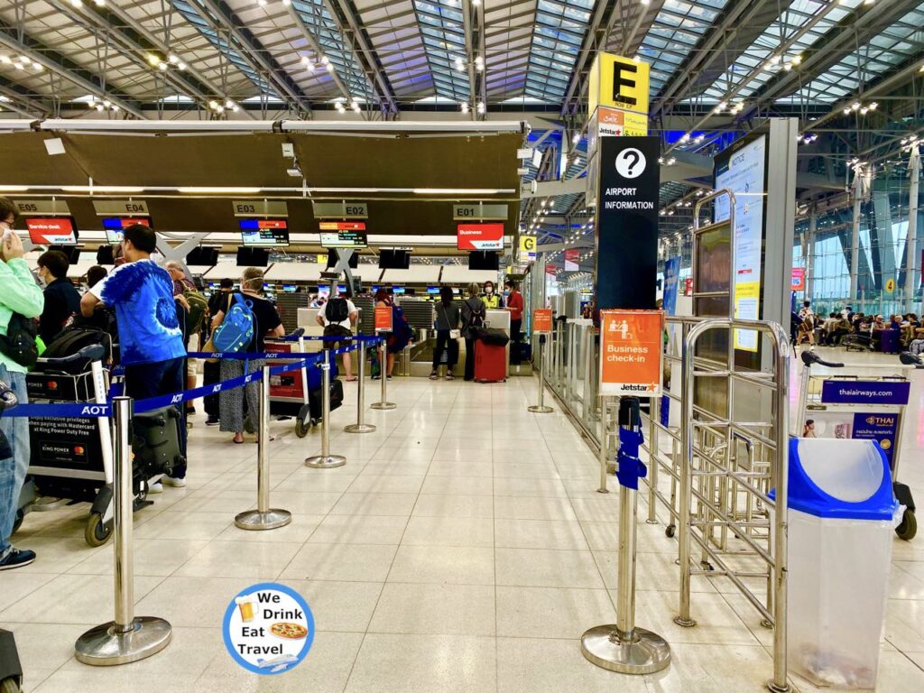 Busy airport check-in counters at Bangkok Airport for JetStar, but with only 2 customers at the business class check-in. People are queuing with luggage, and airport staff are visible behind the counters.
