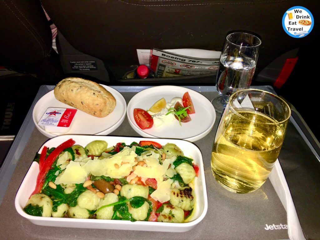 Dinner on board a Jetstar Business Class flight with a long roll with butter, four wedges of tomato on a plate, a plate of gnocchi with vegetables, accompanied by a glass of wine and water.
