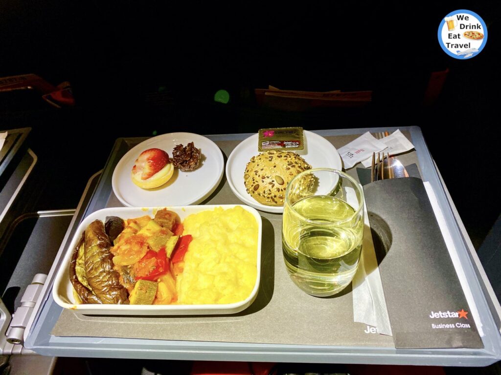 Inflight meal aboard Jestart business class with small plate of fruit, a seeded roll with butter, a plate of vegetables with polenta and a glass of wine with accompanying cutlery and serviettes.