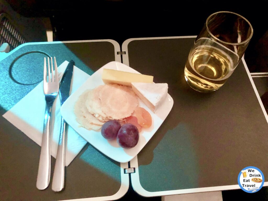 Inflight snack aboard a Jetstar business class flight with a small plate of 2 red grapes, 2 dried apricots, a wedge of soft cheese, a finger of hard cheese and some water crackers, accompanied by a glass of white wine and cutlery.