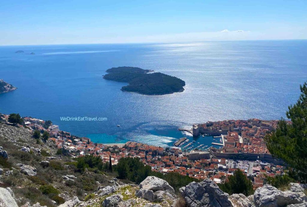 Scenic view of Dubrovnik, Croatia, from Mount Srđ, showcasing Lokrum Island in the centre, the terracotta rooftops of the Old Town, and the Adriatic Sea, under a brilliant blue sky.