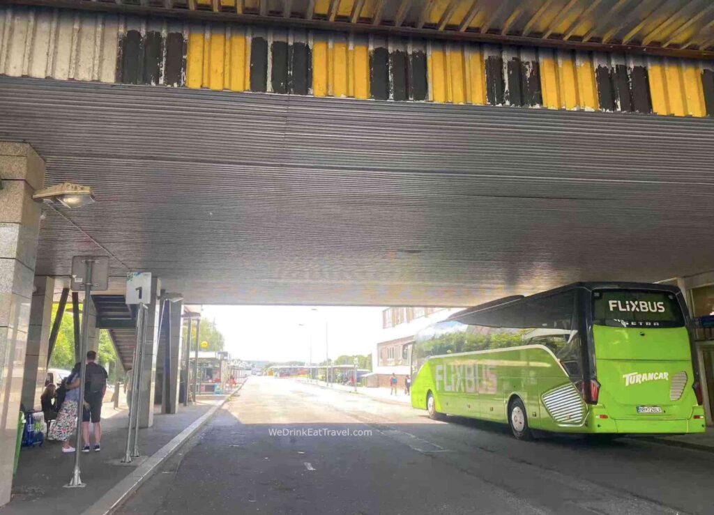 Karlovy Vary Terminál Bus Station with a bright fluorescent colored green Flixbus stationed beneath an overpass with pedestrians walking past.