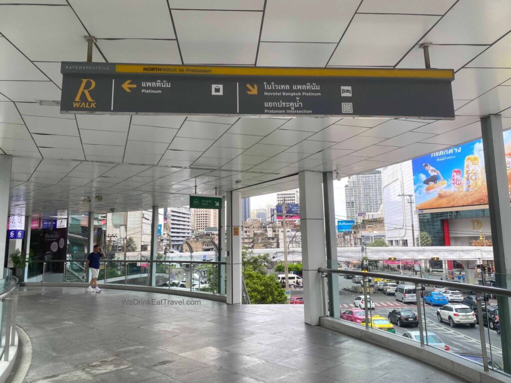 Overhead signage at Ratchaprasong intersection in Bangkok guides pedestrians on how to go to Platinum Fashion Mall, with a busy street scene and city buildings visible below.