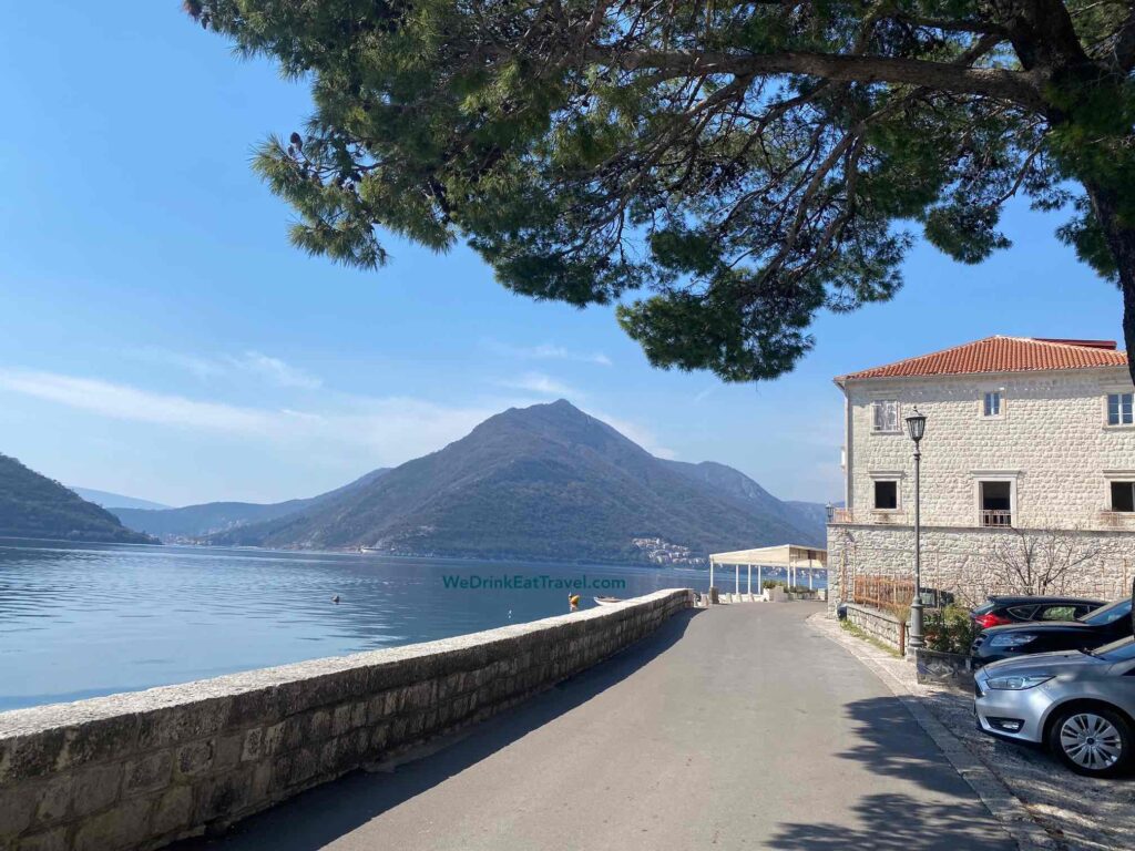A scenic coastal road in Perast, Montenegro, with a stone wall bordering the calm blue bay and a large mountain in the background. A traditional stone building with a red tile roof stands to the right, with parked cars on the street.