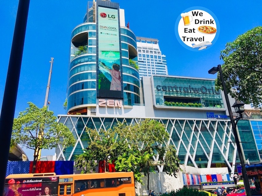 The outside of a CentralWorld shopping centre in Bangkok under a clear blue sky with busy traffic passing by in front.