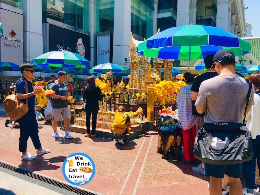People worshipping at the Erawan Shrine in Bangkok, Thailand with blue and green umbrellas set up for shade and shopping centre in the background.